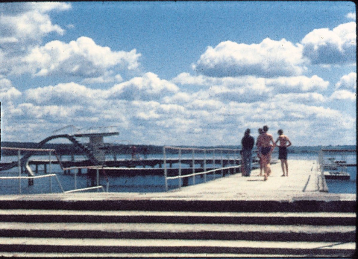 Foto eines Stegs, auf dem sich vier Personen, davon zwei in Badehosen befinden. Linkerhand gibt es eine Rutsche ins Wasser. Weiße Wolken sind am sonst blauen Himmel.