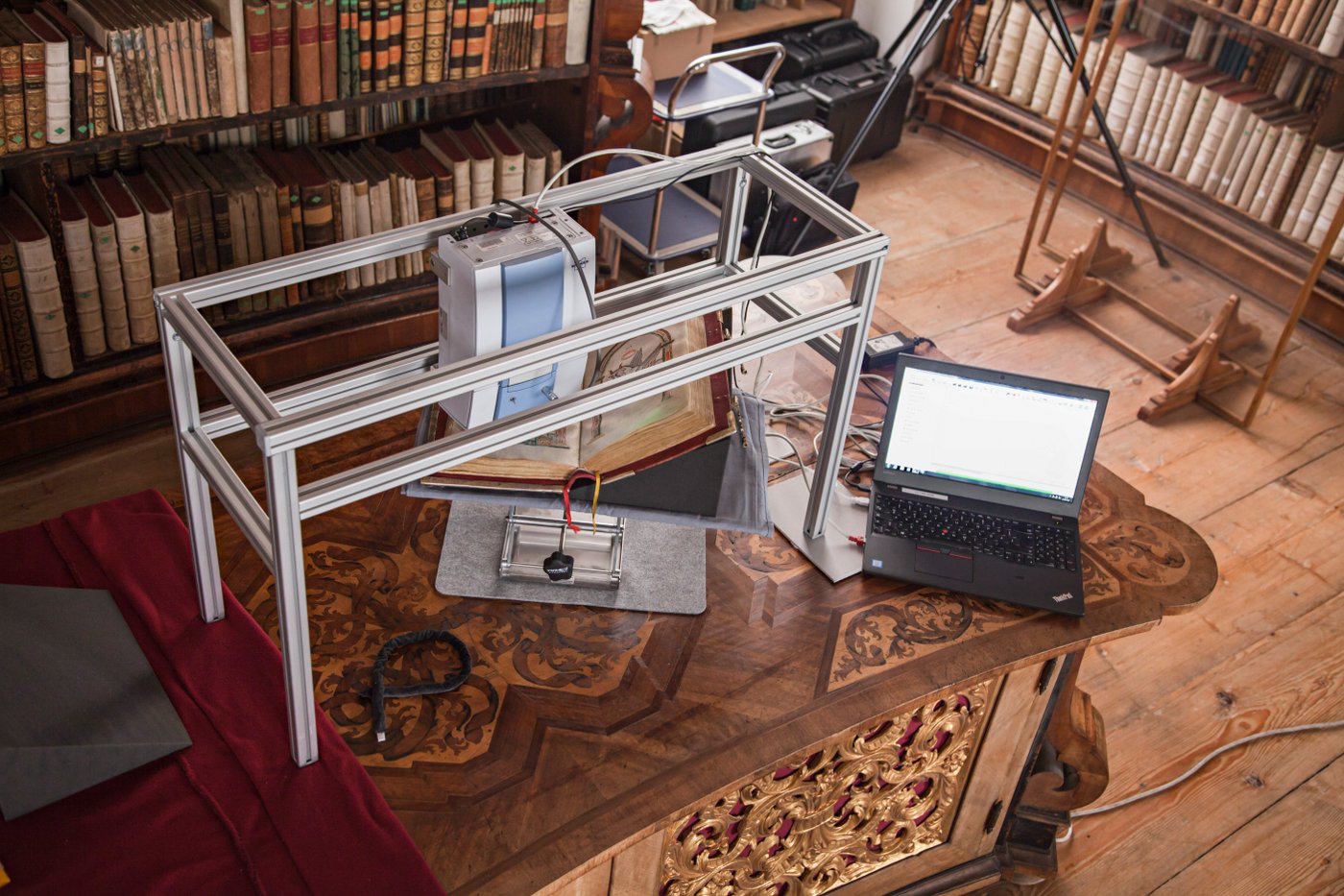 Infrarotspektroskopische Untersuchung einer illuminierten Handschrift in der Bibliothek im Stift Kremsmünster. Regale mit Büchern im Hintergrund, Holzboden und ein Tisch, auf dem ein Laptop sowie die Handschrift und das Untersuchungsgerät stehen.
