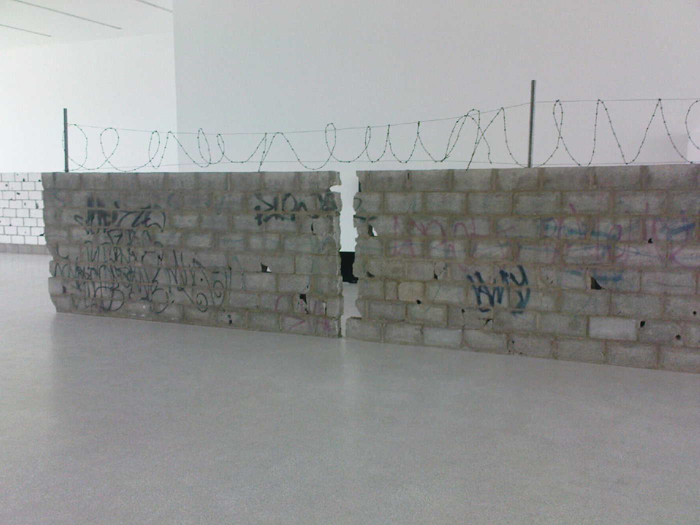 Eine in der Mitte gespaltene Mauer in einer Ausstellung. Die grauen Ziegel sind sichtbar, über die gesamte Mauer verläuft ein Stacheldrahtzaun.