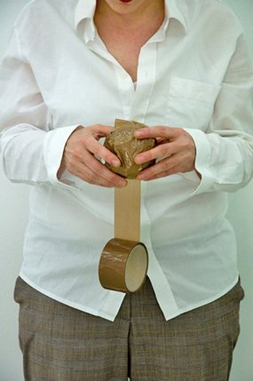Vor dem in eine weiße Bluse gehüllten Oberkörper hält die Person mit ihren Händen eine braune Kugel, daran heftend eine braune Klebebandrolle, die ca. 20 Zentimeter entrollt ist.
