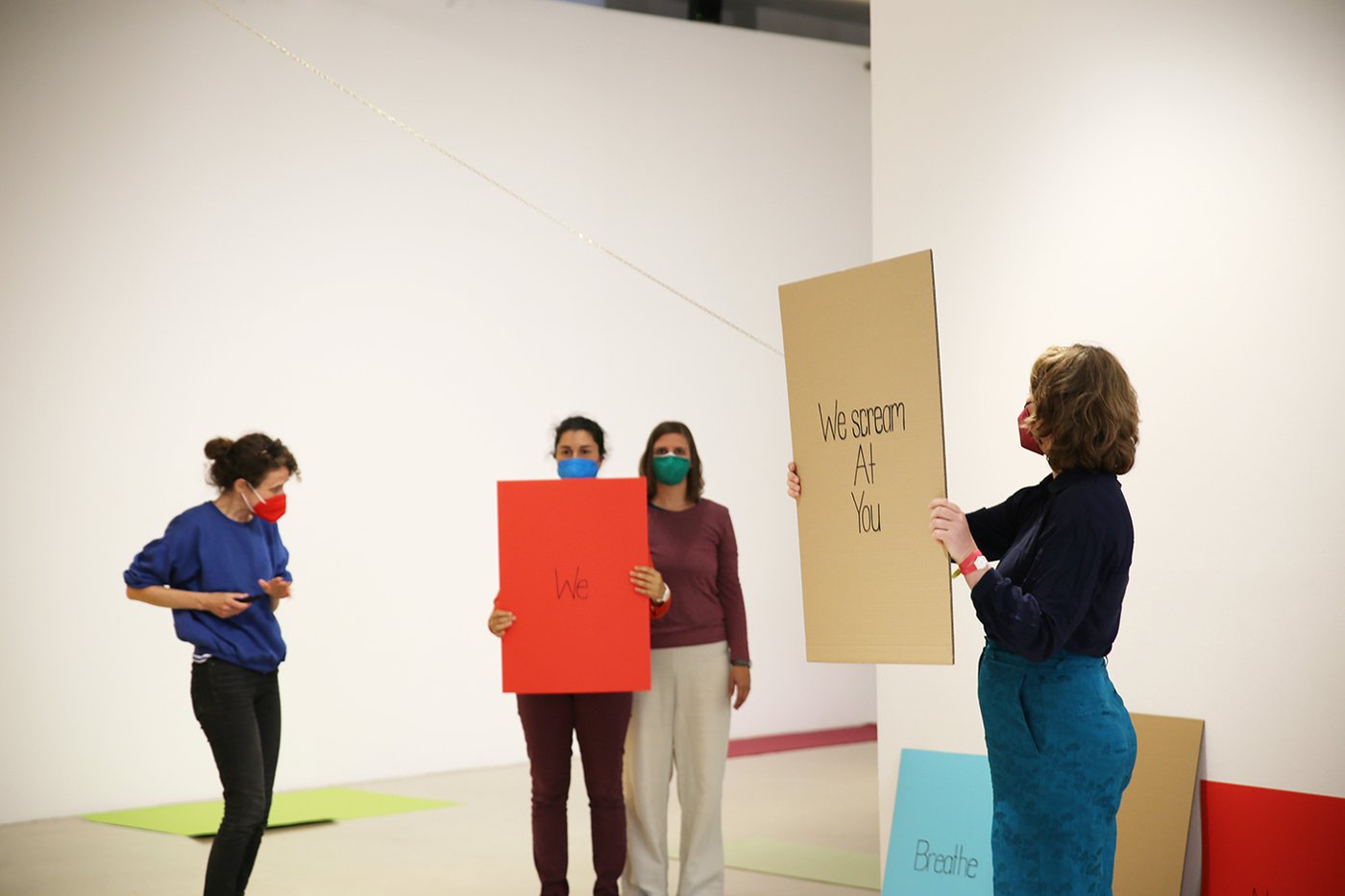 Vier Frauen, die Masken tragen. Zwei davon halten Schilder mit Text hoch, "We" und "We scream at you".