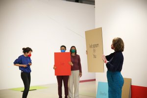 Vier Frauen, die Masken tragen. Zwei davon halten Schilder mit Text hoch, "We" und "We scream at you".
