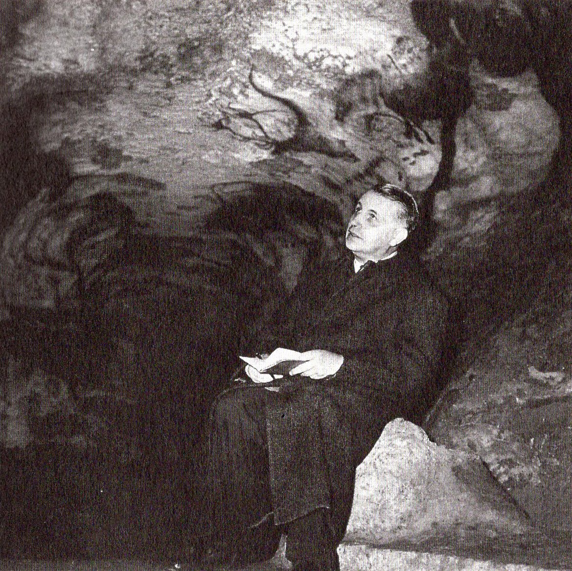 Georges Bataille sitzt mit einem Notizblock in der Höhle von Lascaux und blickt auf die Decke.