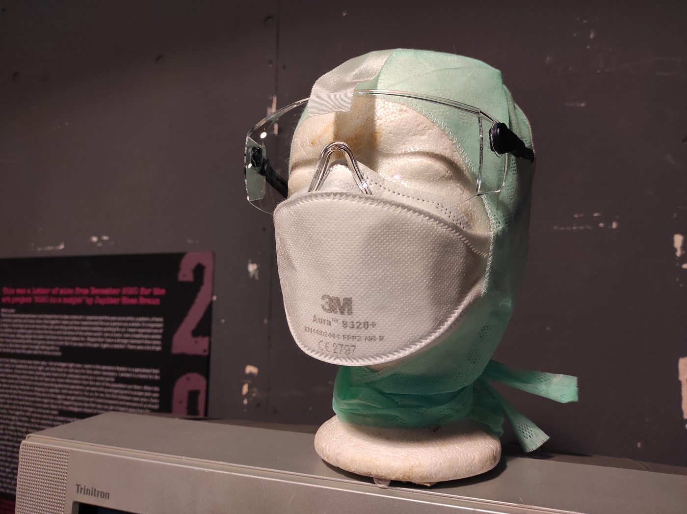 Eine Styropor-Kopf mit FFP2 Maske, Schutzbrille und Kopfschutz steht auf einem Triniton-Monitor.