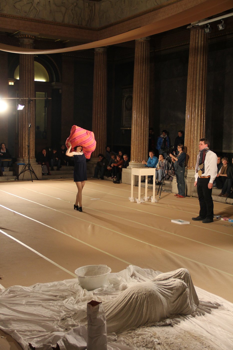 Bild2 A performer crosses the auditorium with a red fabric structure on her head. In the foreground a performer lies veiled on the floor