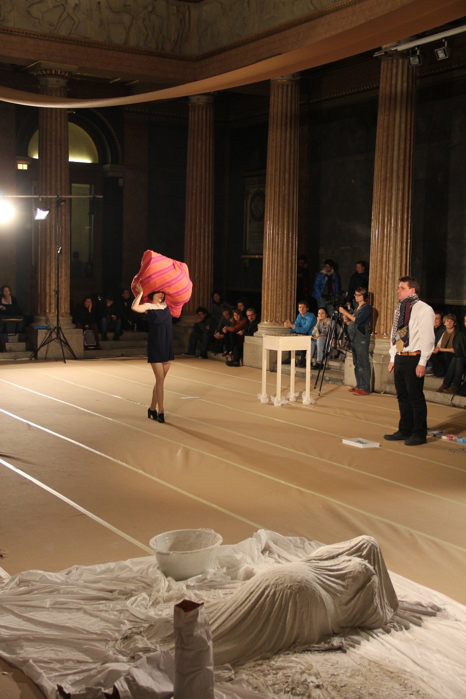 A performer crosses the auditorium with a red fabric structure on her head. In the foreground a performer lies veiled on the floor