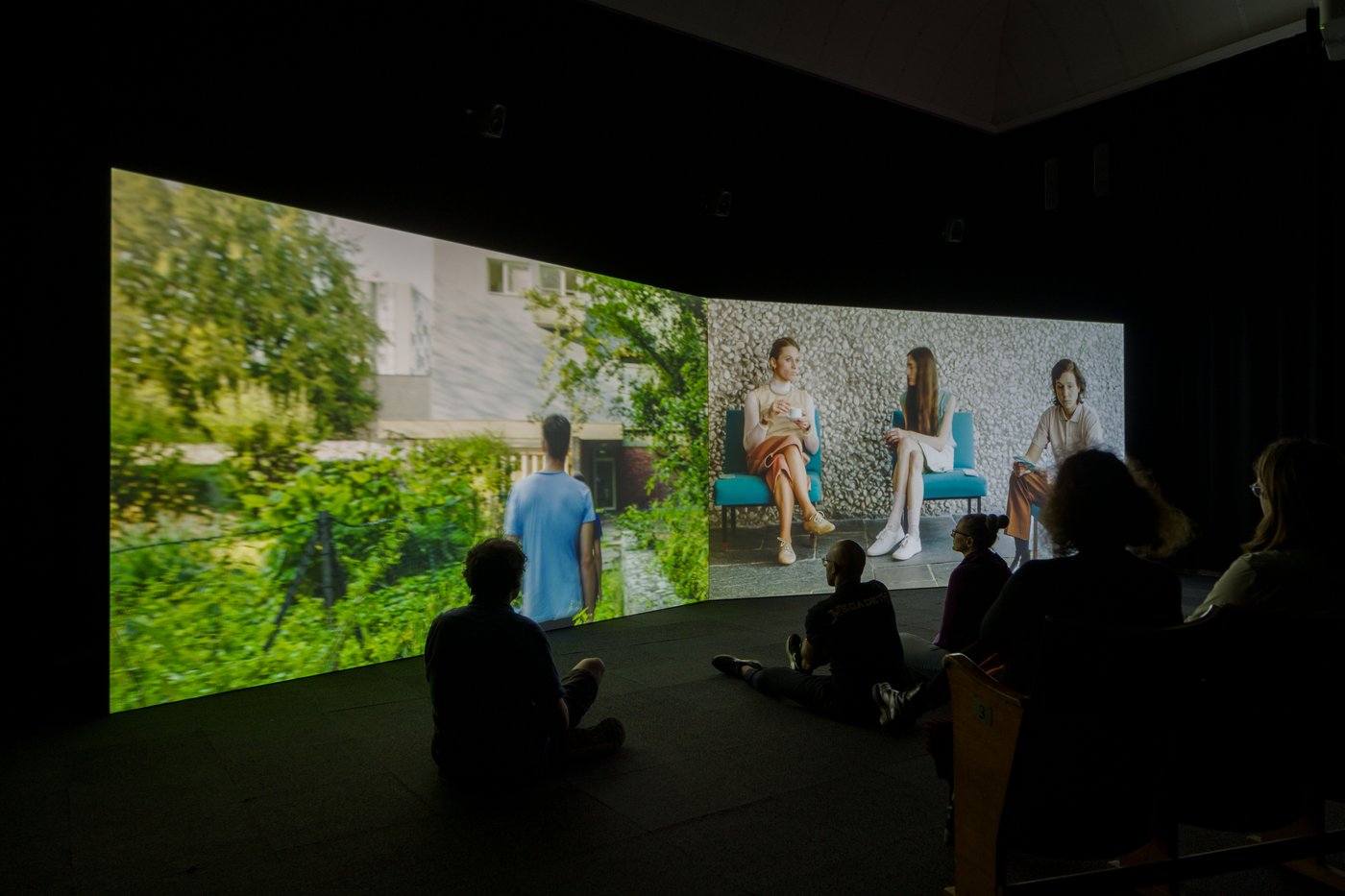 2 2-part screen, one with a man in a garden, another with three young women on blue armchairs. In front of it people are sitting in the dark.