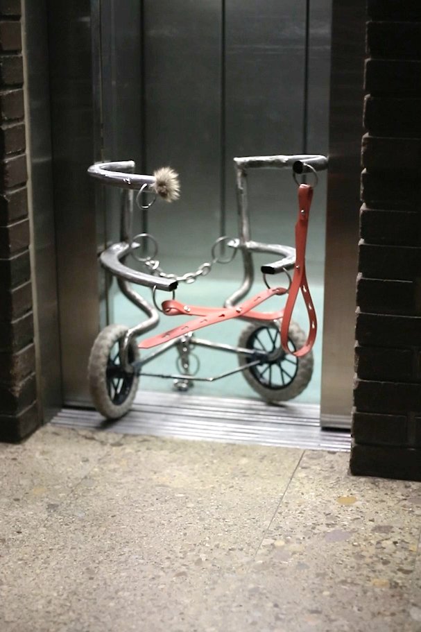 Foto von einem fetischartigen Gefährt mit 2 Rädern, Ledergürteln und Ketten im Eingang eines Aufzugs.