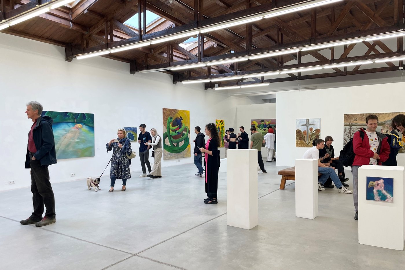 Besucher_innen tauschen sich über die Werke in der Galerie aus. An den drei Wänden und drei im Raum stehenden Sockeln sind 8 Gemälde zu sehen.