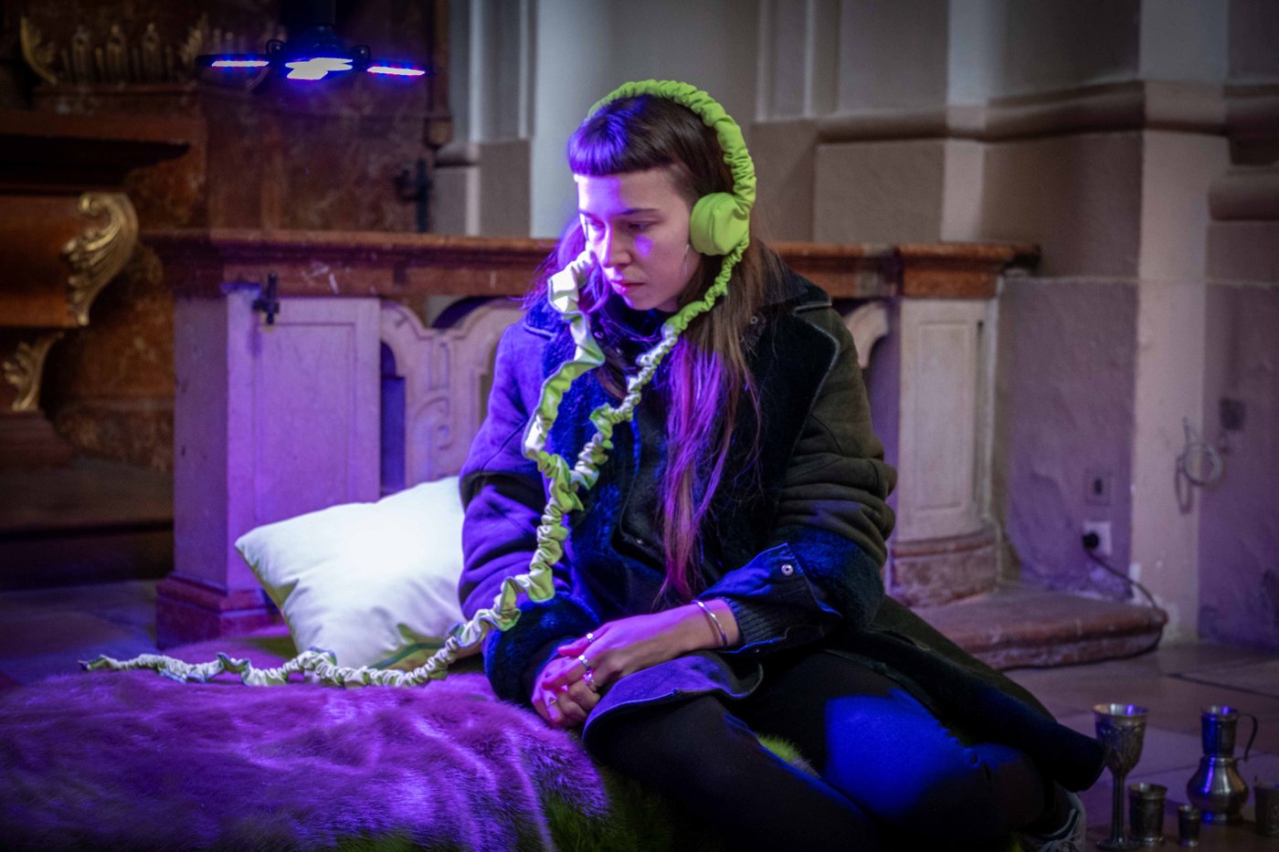 Frau mit grünen Kopfhörern sitzt auf einer Matratze in einer Kirche und wird von blauem Licht beleuchtet