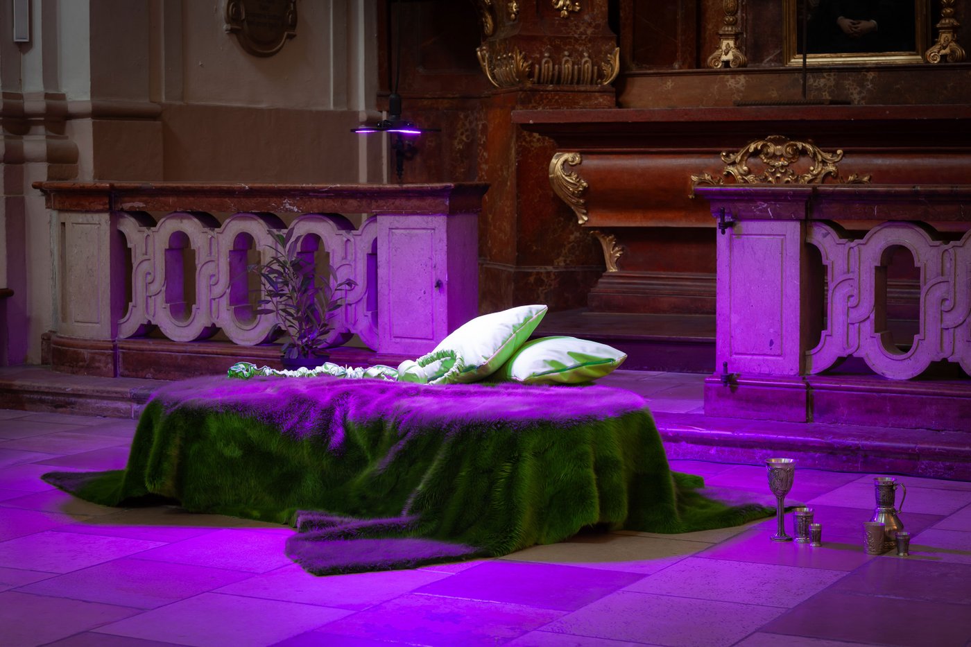 Grüne Matratze vor einem Kirchenaltar, die mit rosa Licht beleuchtet wird mit Kopfhörern, am Boden mehrere Krale