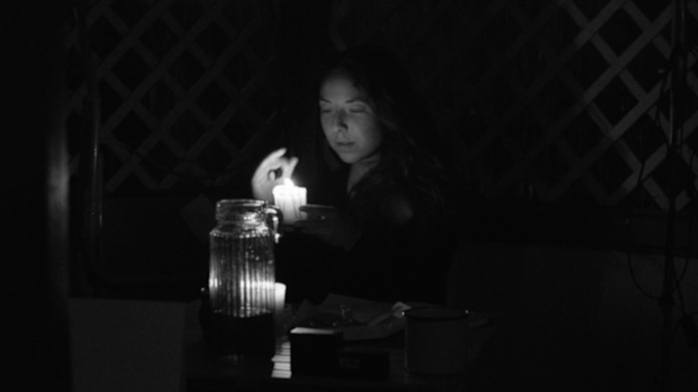Ein Filmstil vom Film “Eine Begegnung”. Eine Frau zündet eine kleine Kerze im Dunkel an. Sie schaut das Licht einer Kerze.