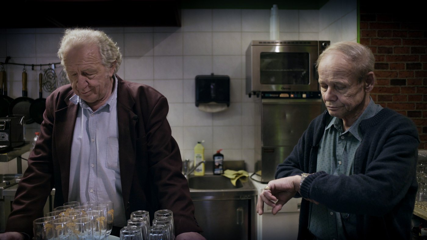 zwei ältere Männer in einer Küche, rechts der Mann schaut auf seine Uhr, der mann links hält ein Tablett mit Gläsern