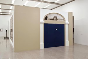 Ansicht eines Ausstellungsraumen, in der Mitte eine blaue geschlossene Türe umrahmt von weißen Säulen, darüber ein Türbogen mit einer Radierung darin, beige Wände auf den Seiten der Türe, links hängt ein Kunstwerk an der anderen Wandseite und im Hintergrund ist ein Bildschirm zu sehen.