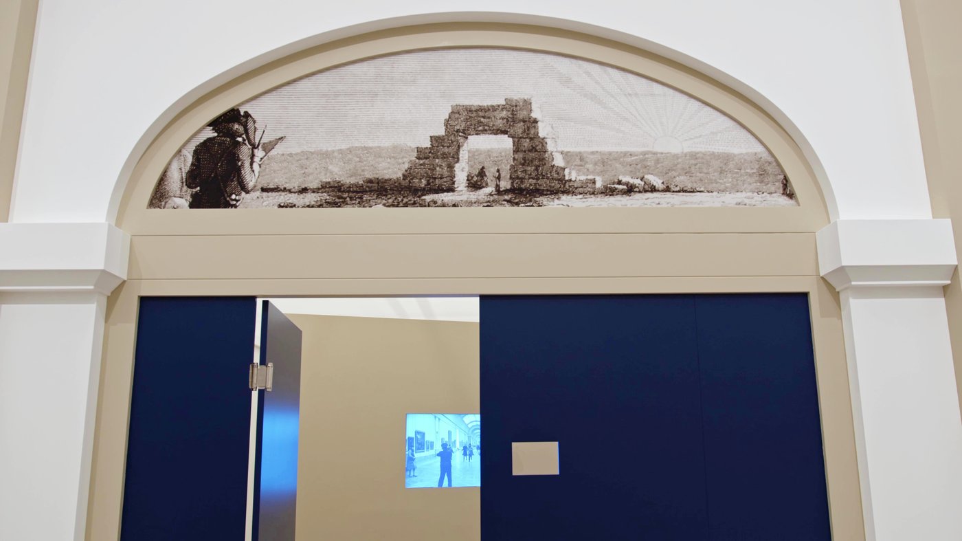 Fotografie einer blauen Doppeltüre, bei der eine Seite offen ist und Blick auf einen Bildschirm im Hintergrund freigibt, darüber ein Türbogen in dem ein Ausschnitt einer Radierung in der ein Sonnenaufgang und eine Steinruine und Personen zu sehen sind