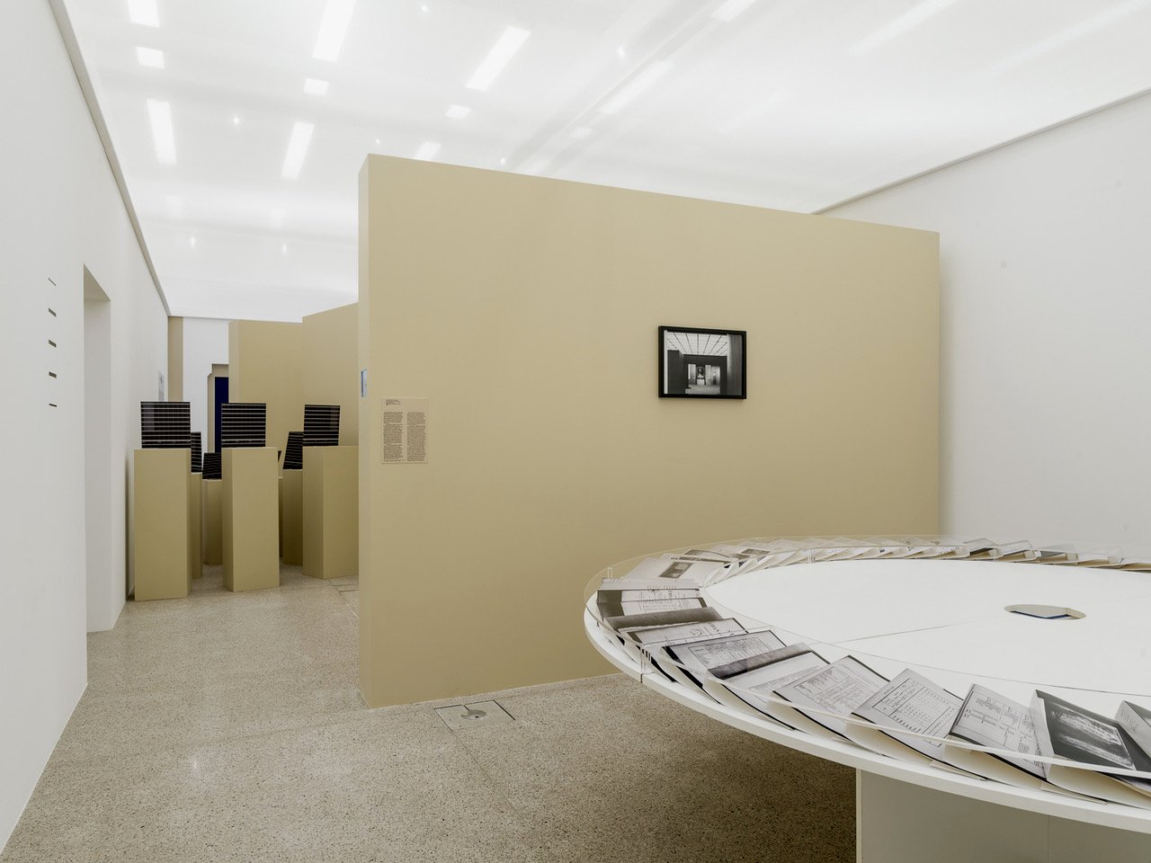 Fotografie eines Ausstellungsraumes mit beigen Trennwänden auf denen Kunstwerke plaziert sind, im Vordergrund ein runder weißer Tisch mit Mappen die im Kreis aufgelegt wurden ,links hinten schwarze Gegenstände auf Podesten