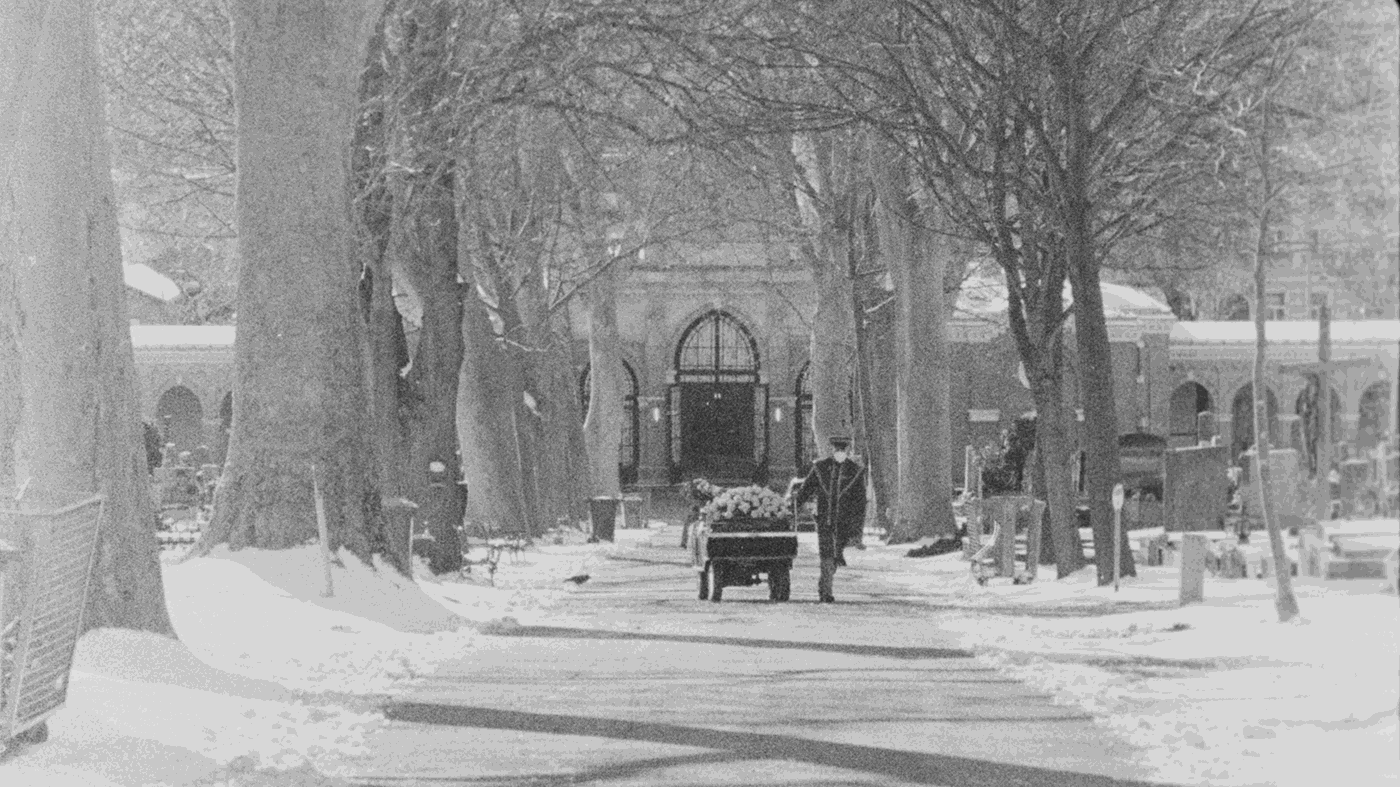 schwarz-weiß Aufnahme von einem Friedhofsangestellten der einen mit Blumen geschmückten Sarg eine Allee am Friedhof, der mit Schnee bedeckt ist, entlang fährt