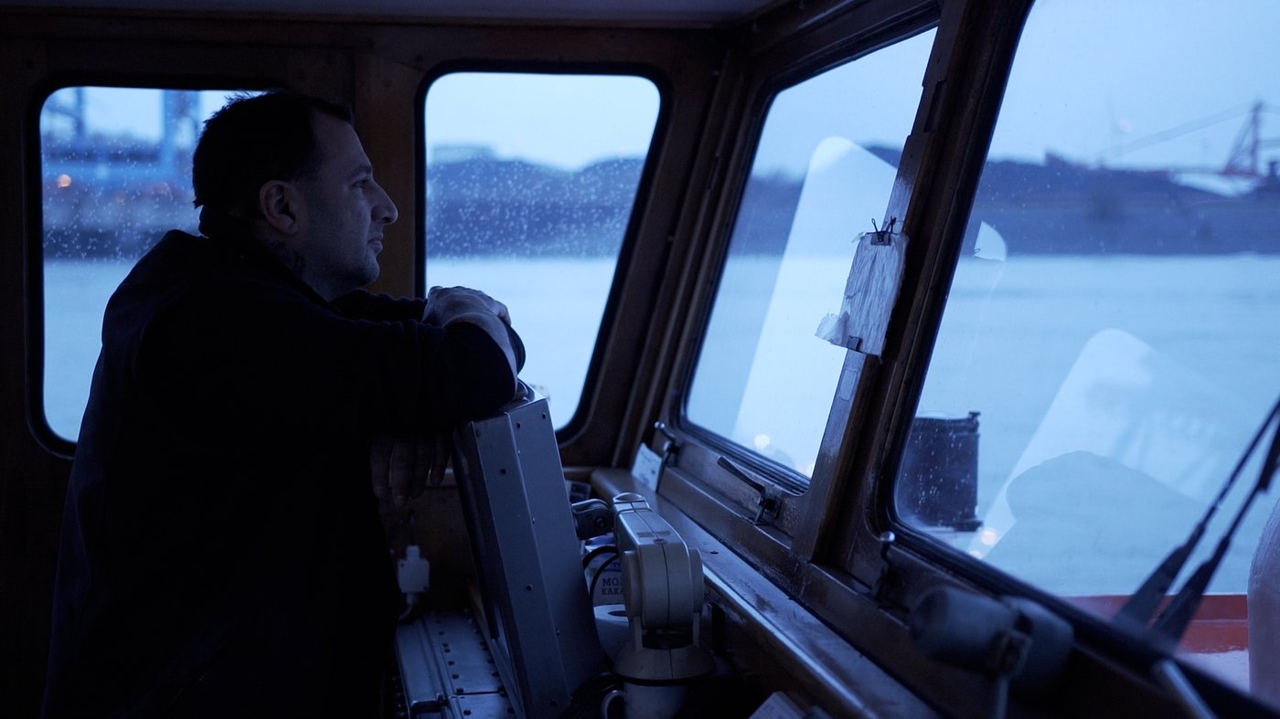 Foto eines Mannes der im Schiffskontrollraum steht und auf seinen Instrumenten lehnt und aus dem Fenster blickt