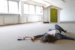 Person liegt in einem hellen Raum am Boden als wäre sie gerade gestürzt, mit offener Tasche neben ihr am Boden, im Hintergrund ein kleines Holzhaus mit Puppe am Dach