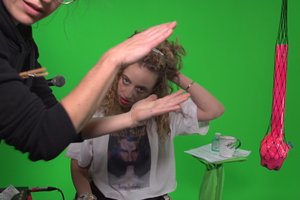 Die Abbildung zeigt ein Standbild aus dem Video Green Scream mit G-udit, im Vordergrund klatscht eine Hand, im Hintergrund eine Frau fährt sich eine Frau mit ihrer Hand durch die Haare.