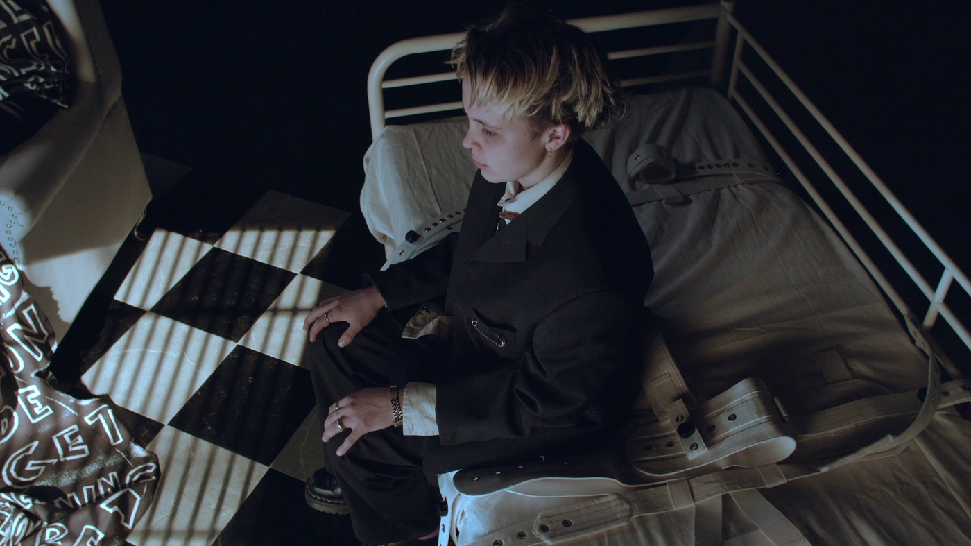 Videostill: Perspektive von schräg oben. Eine junge Person in einem schwarzen Anzug sitzt aufrecht auf einem Bett mit weißer Matratze und weißem Metallrahmen. Links und Rechts neben der Person liegen weiße Fixiergurte aus Leder. Der Boden ist in einem schwarz-weißen Schachbrettmuster gekachelt und führt ins Dunkle. Auf den Boden fällt der Schatten von aufgezogenen Fensterlamellen.