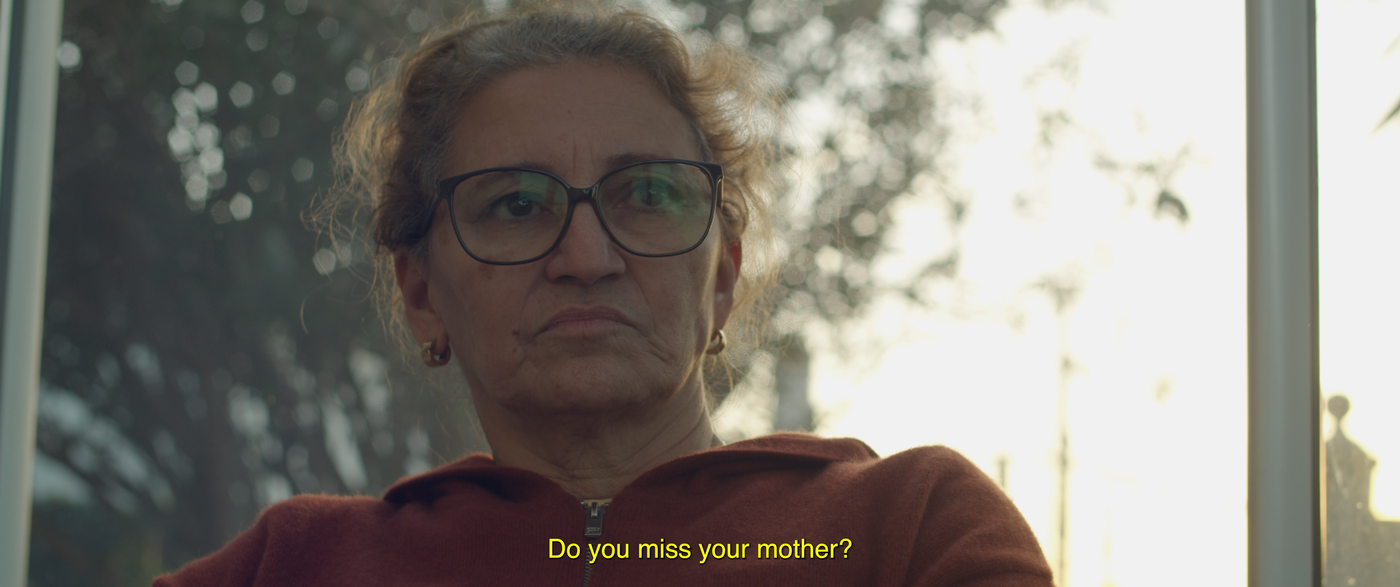 Videostill. Das Porträt einer älteren Frau mit strengem Blick, vor einem Fenster. Im Hintergrund eine verschwommene Baumkrone. Am unteren Bildrand sind englische Untertitel eingeblendet: „Do you miss your Mother?“