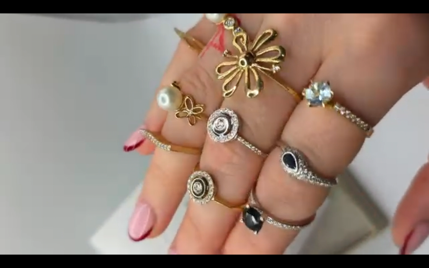 Videostill. Ausschnitt einer Hand mit vielen extravaganten Ringen vor hellem Hintergrund. Altrosa Nägel mit dunkelroten Nagelspitzen.