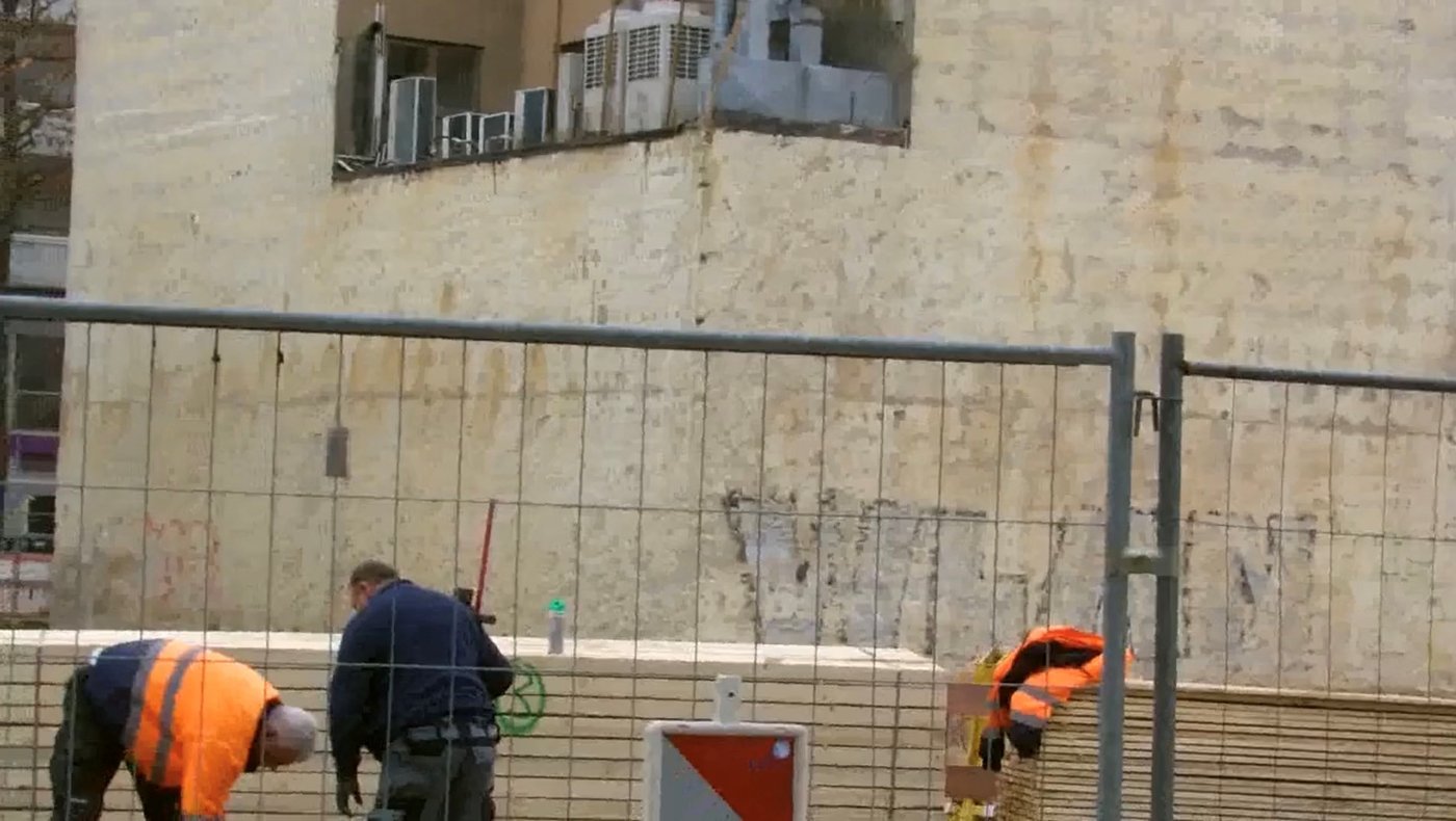 Videostill. Drei Bauarbeiter, einer dunkel gekleidet, zwei in orangenen Warnjacken, arbeiten an einer Baustelle vor einer kargen Hauswand. Im Bildvordergrund ist ein metallenes Baugitter.