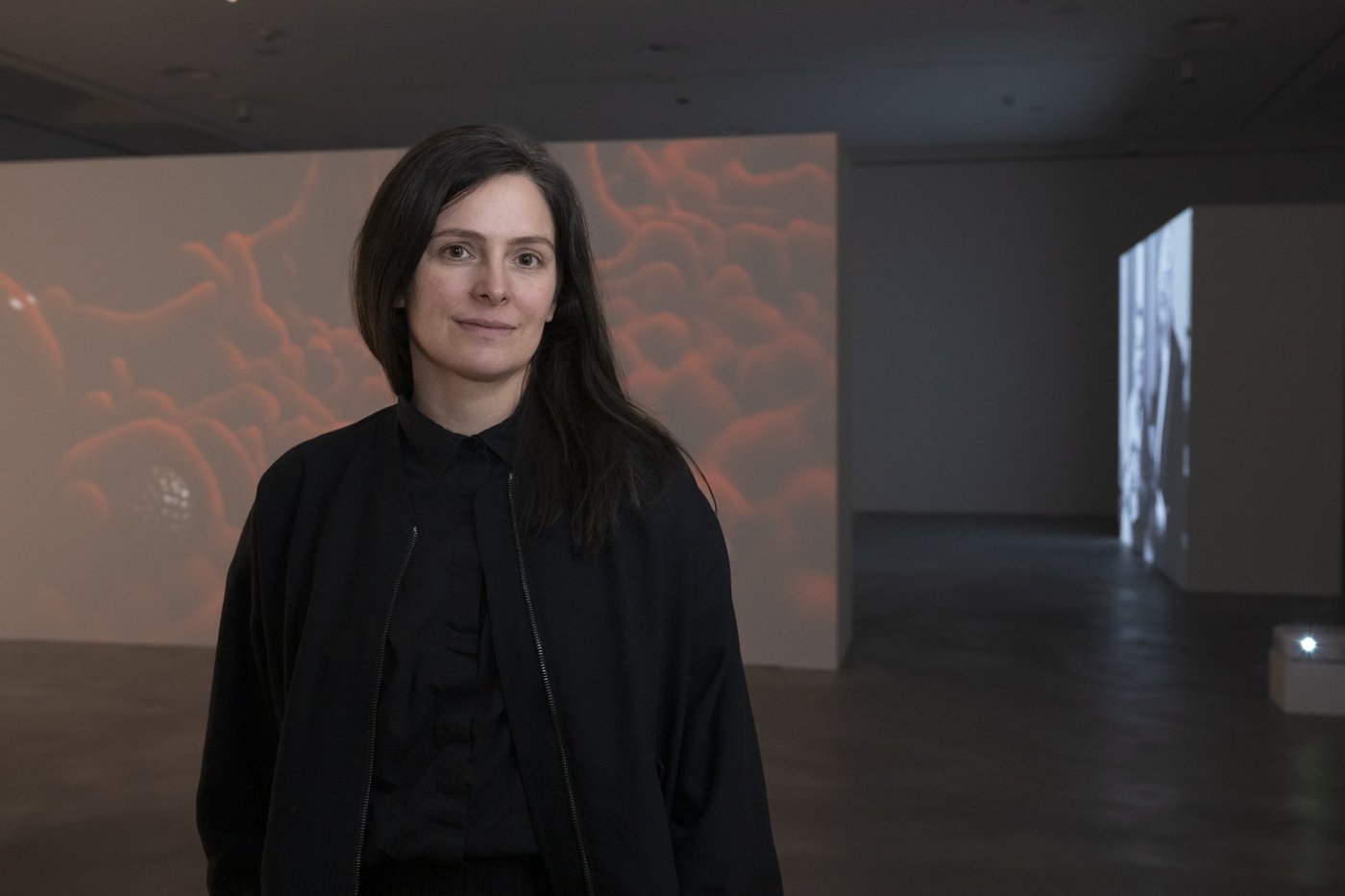 Porträt von Claudia Larcher in schwarzer Kleidung in einem dunklen Ausstellungsraum, hinter ihr eine Wandprojektion mit rötlichen, wolkenartigen Formen.