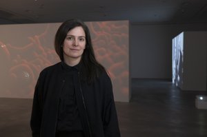Porträt von Claudia Larcher in schwarzer Kleidung in einem dunklen Ausstellungsraum, hinter ihr eine Wandprojektion mit rötlichen, wolkenartigen Formen.