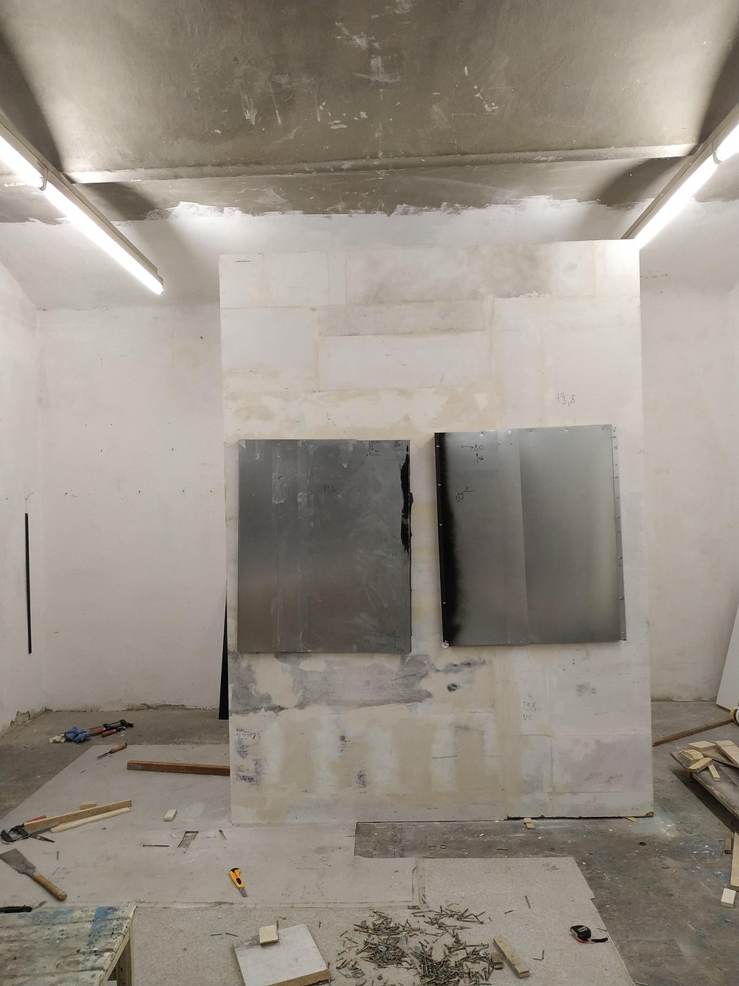 Ein Atelierraum mit freistehend gezimmerter Wand in der Mitte, auf der 2 große dunkelgrau/silberne flache Blechobjekte angebracht sind. Die Objekte sind fast so breit wie die Wand und hängen etwas schief. An den Rändern sind die Blechtafeln teilweise schwarz eingefärbt. Am Boden liegen Werkzeuge, Schrauben, Pappen und Holz