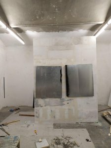Ein Atelierraum mit freistehend gezimmerter Wand in der Mitte, auf der 2 große dunkelgrau/silberne flache Blechobjekte angebracht sind. Die Objekte sind fast so breit wie die Wand und hängen etwas schief. An den Rändern sind die Blechtafeln teilweise schwarz eingefärbt. Am Boden liegen Werkzeuge, Schrauben, Pappen und Holz