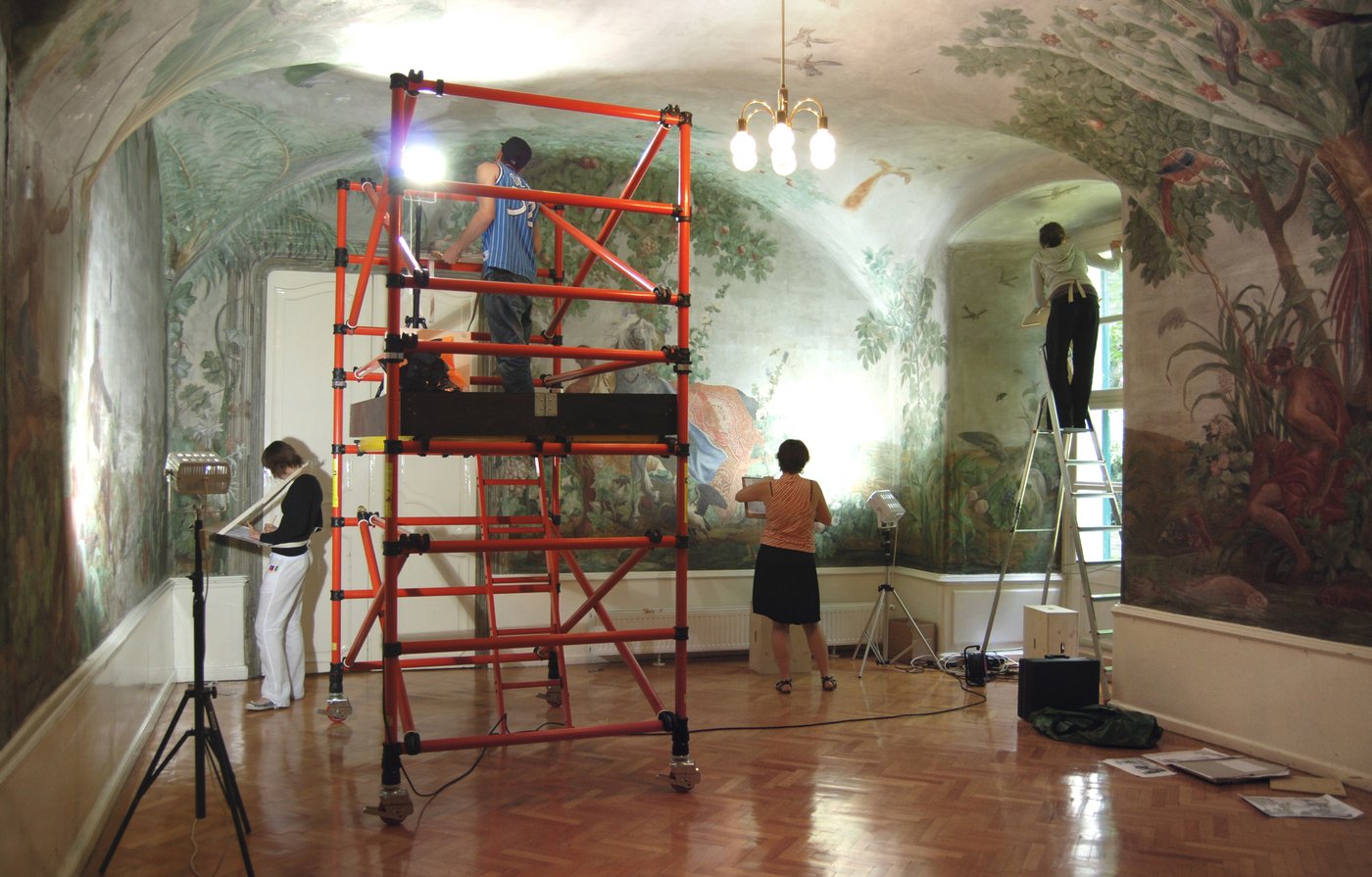 Four students working on the walls of the room, one is standing on a scaffolding.