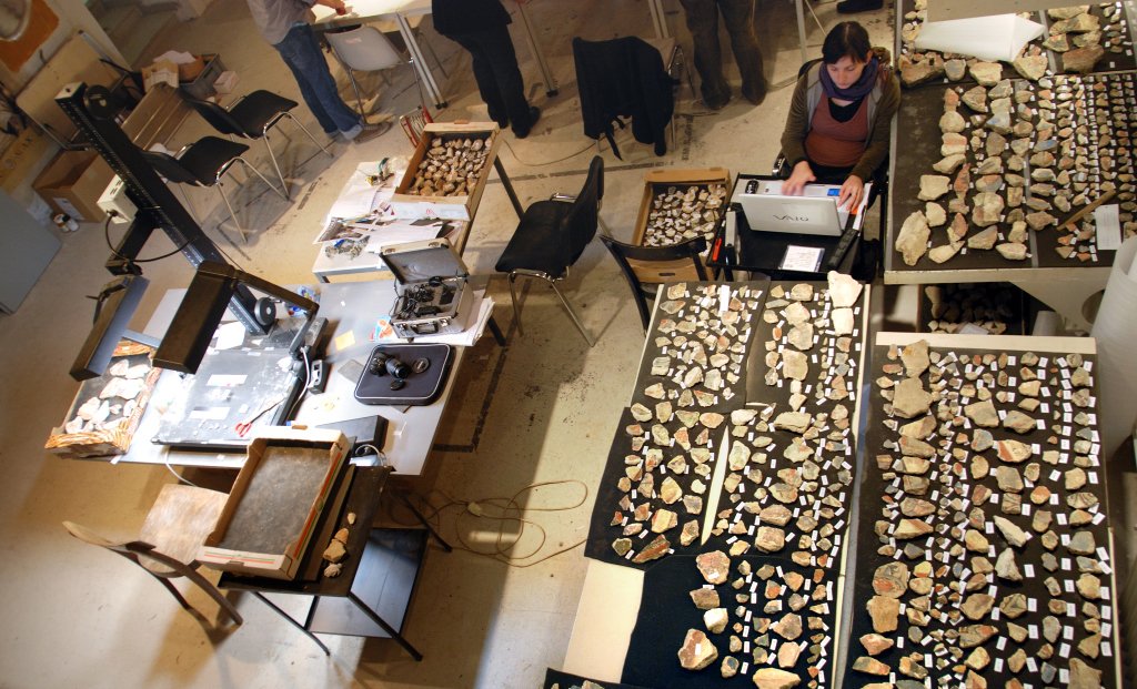 Numerous fragments are carefully lined up on tables, one student is sitting behind a computer.