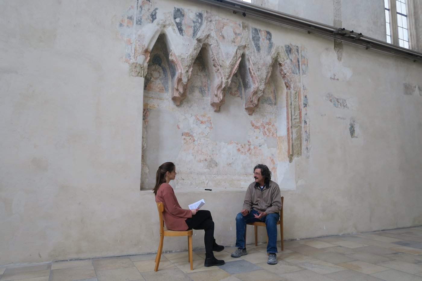 Eine Forscherin interviewt einen Restaurator vor einer historischen Steinmauer