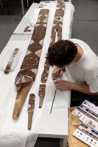 eine Person sitzt an einem Tisch auf dem eine große Holzmaske liegt, die er mit feinem Werkzeug restauriert, Foto aus der Vogelperspektive