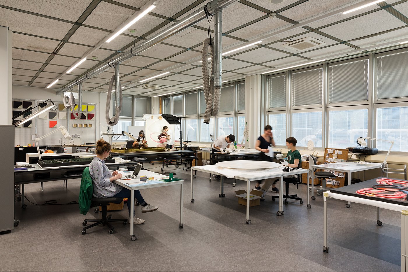 Ansicht eines Atelieraumes mit Personen die an ihren Tischen sitzen und mit technischen Geräten an Werken arbeiten