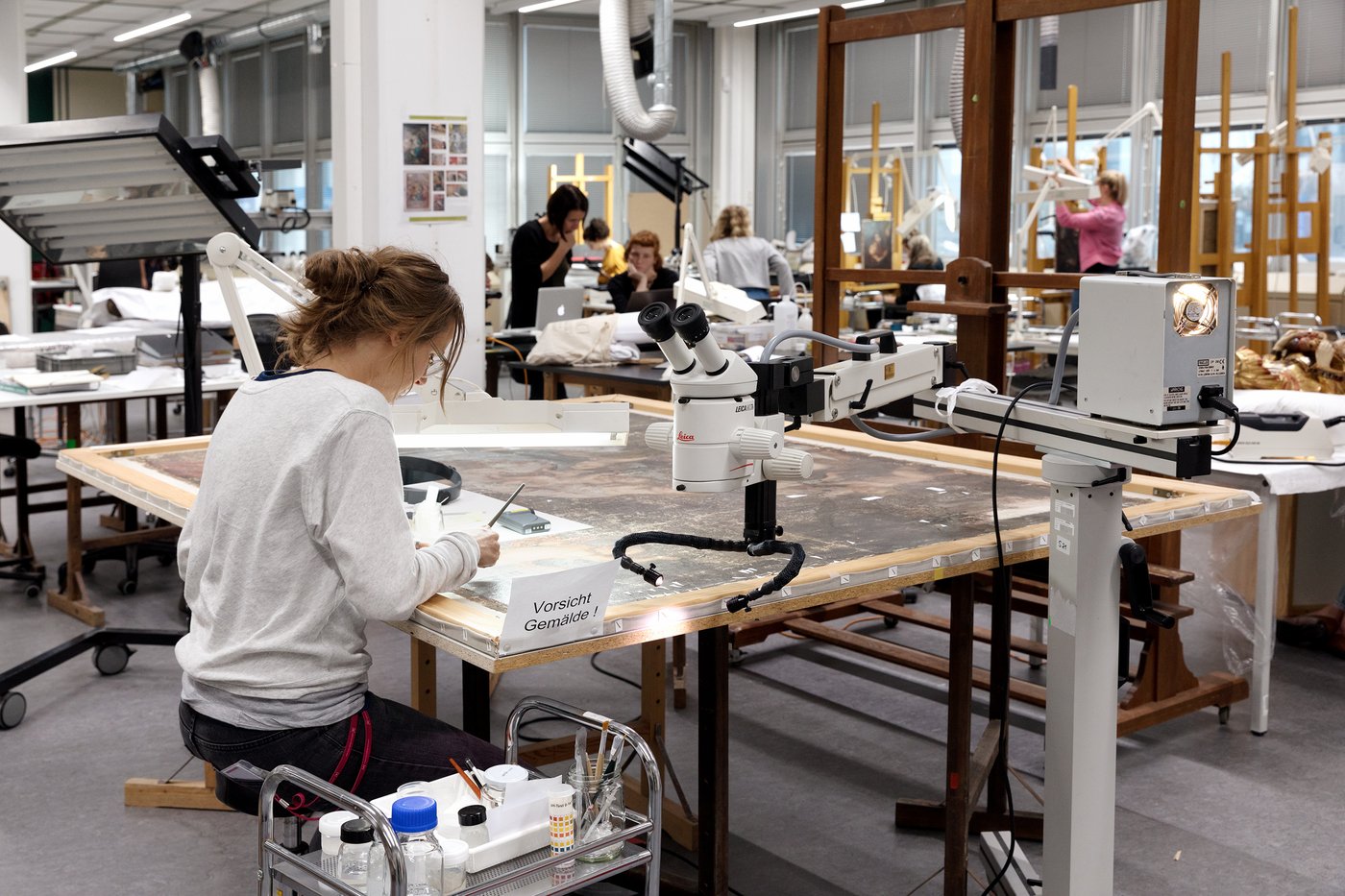 Blick in einen großen, hellen Atelierraum mit Fensterfront rechts. Im Vordergrund arbeitet eine Studentin an einem Gemälde, das auf dem Tisch liegt. Im Hintergrund sind einige Studierende und Staffeleien zu sehen.