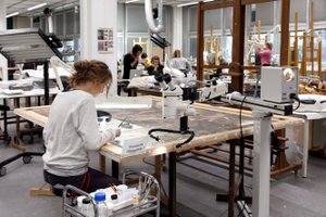 Blick in einen großen, hellen Atelierraum mit Fensterfront rechts. Im Vordergrund arbeitet eine Studentin an einem Gemälde, das auf dem Tisch liegt. Im Hintergrund sind einige Studierende und Staffeleien zu sehen.