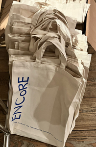Mehrere beigefarbene Stofftaschen mit ENCoRE-Logo liegen gestapelt auf einem Holzboden.