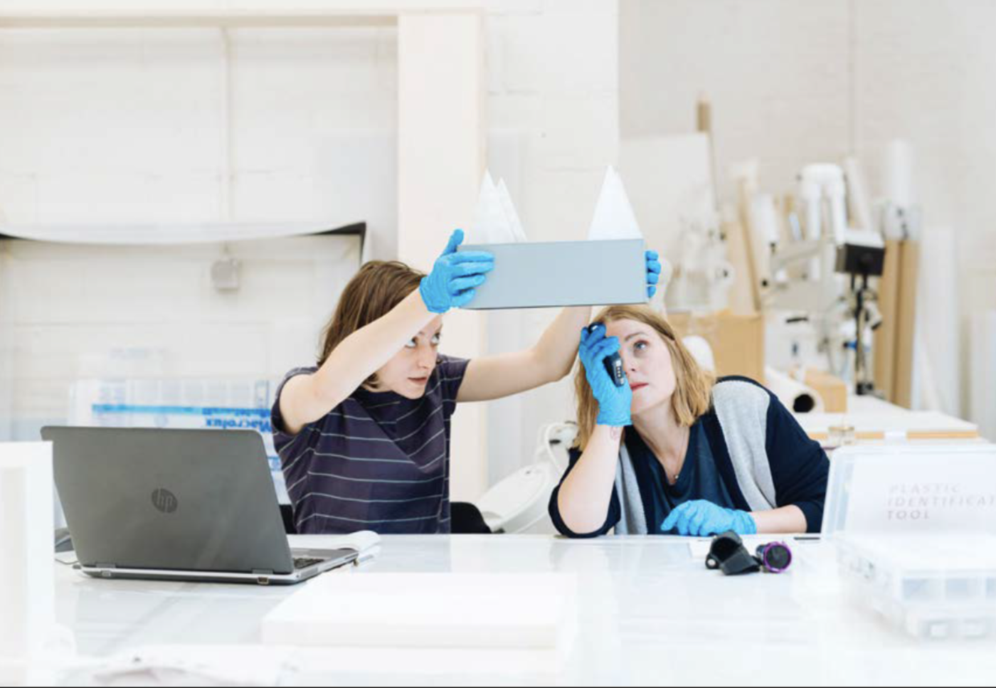 Zwei Personen mit blauen Schutzhandschuhen untersuchen in einem Werkstattraum ein Objekt; eine Person hält eine Probe hoch, die andere betrachtet sie mit einem Handgerät.