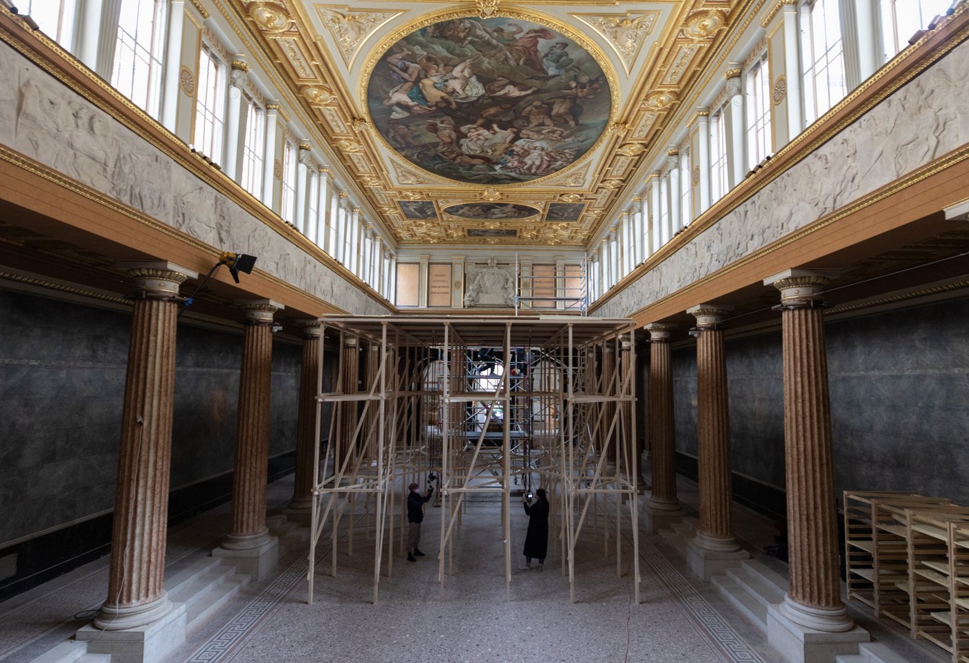 Klassische Architektur der Aula am Schillerplat mit dem Holzgerüsz der Installation und Personen im Raum