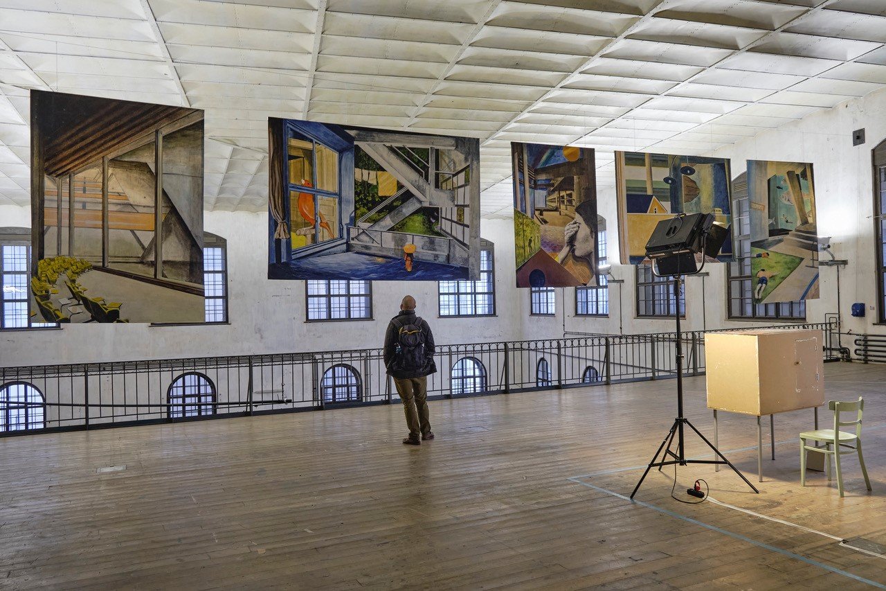heller Ausstellungsraum mit großen Gemälden, die in derLuft hängen, in der Mitte Rückansicht von Mann der sich die Kunstwerke anschaut