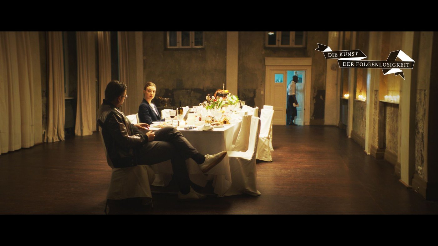 Filmstill mit Blick in einen Saal mit einer Tafel auf der Speisen angerichtet sind und zwei Personen die am Tisch sitzen und miteinander sprechen