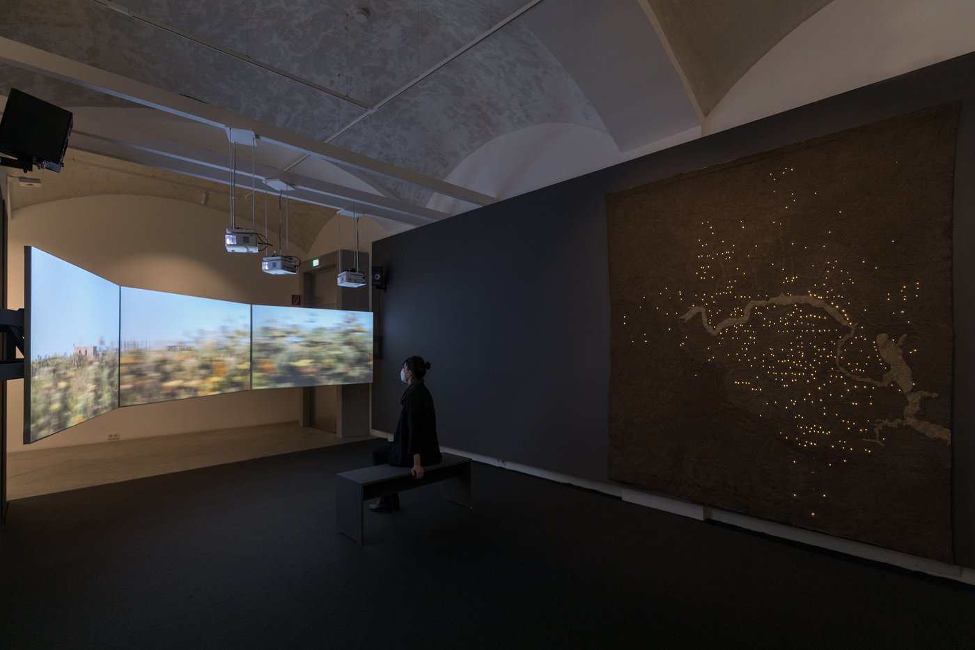 Ausstellungsansicht mit einer Besucherin, zu sehen ist eine Drei-Kanal-Videoinstallation auf der eine vorbeiziehende Landschaft gezeigt wird.