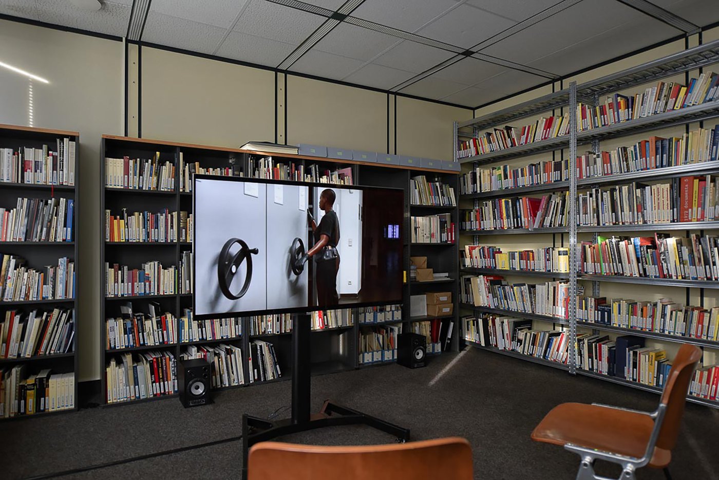 Ansicht der Ausstellung im Archivraum. Im Hintergrund Bücherregale, davor ein freistehender Monitor auf dem ein Video läuft. Auf dem Standbild sieht man eine Person, die bedient das Drehrad eines Archivregals und schaut in den Spalt zwischen zwei Regalen.