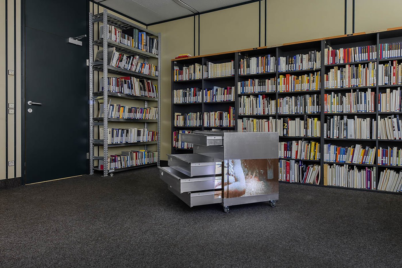 Ansicht der Ausstellung im Archivraum. Im Hintergrund Bücherregale. Davor ein fahrbarere, niedriger silberner Metallkasten mit ausgezogenen Schubladen. Auf die Seite wird ein Film projiziert. Auf dem Standbild erkennt man eine Person mit vor der Brust überkreuzten Armen.