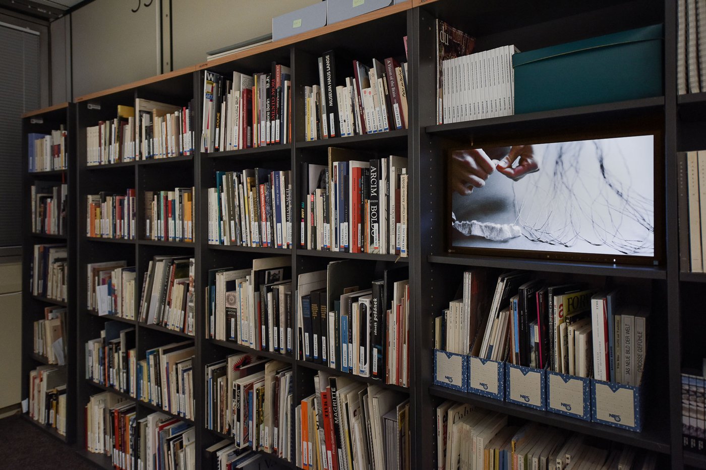 Bücherregal, schräg fotografiert, in einem der Fächer ist ein Monitor zu sehen, auf dem ein Video läuft. Darauf sind Hände zu sehen, die einen Papierfaden knüpfen.
