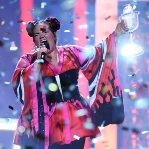 Foto einer Frau in bunter Kleidung die mit Mikrofon auf einer Bühne steht und hineinschreit mit einem Award in ihrer linken Hand und Konfetti um sie herum.