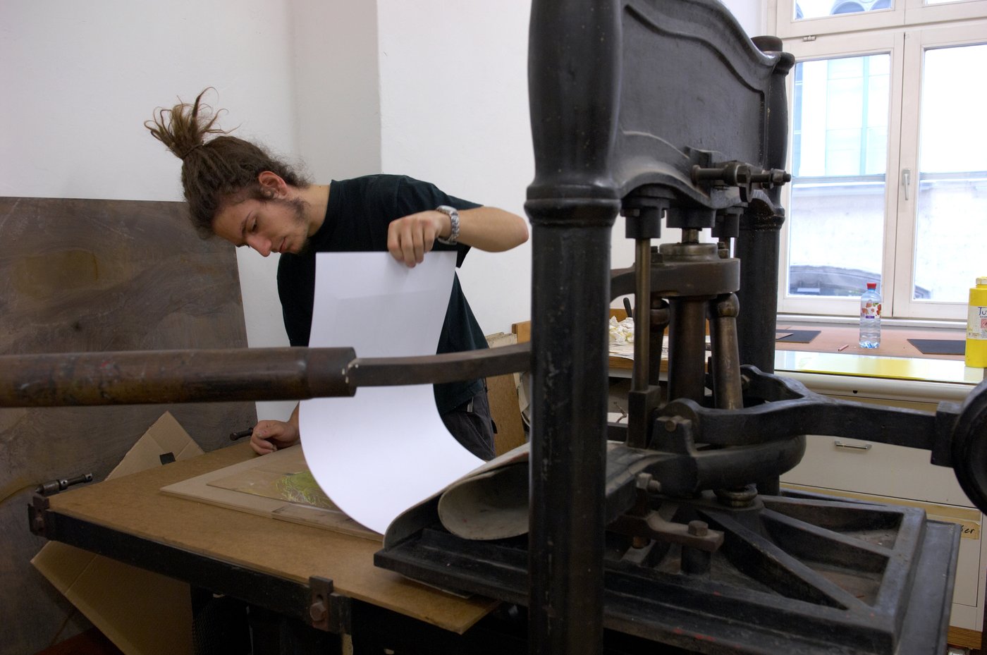 Ein Student steht an einer großen schweren Druckermaschine und überprüft gerade das Ergebnis, indem er das Falzblatt hochhebt.